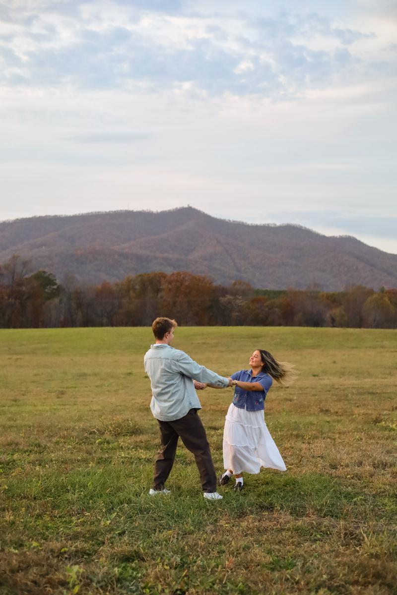 A couple holding both hands and spinning in an open field, a long wooded ridge behind them in the last of the evening light