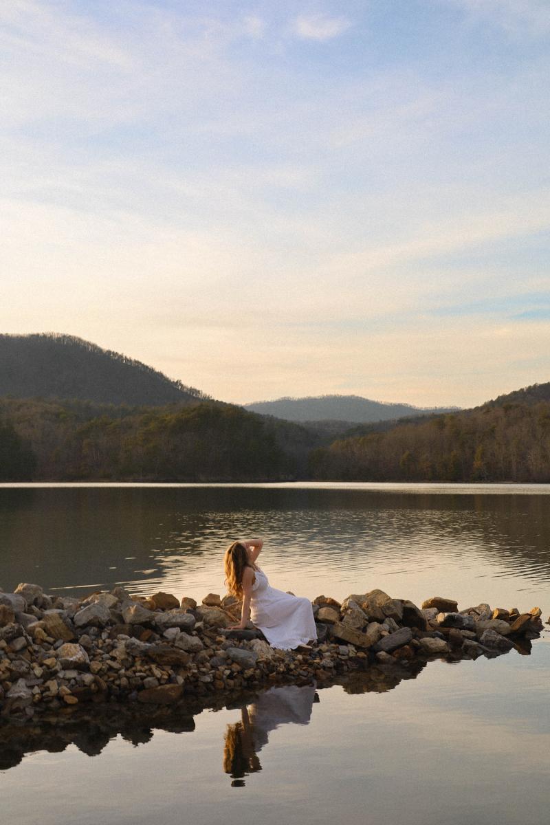 A woman in a long white dress sitting on a rocky shoreline, still water and low wooded hills behind her