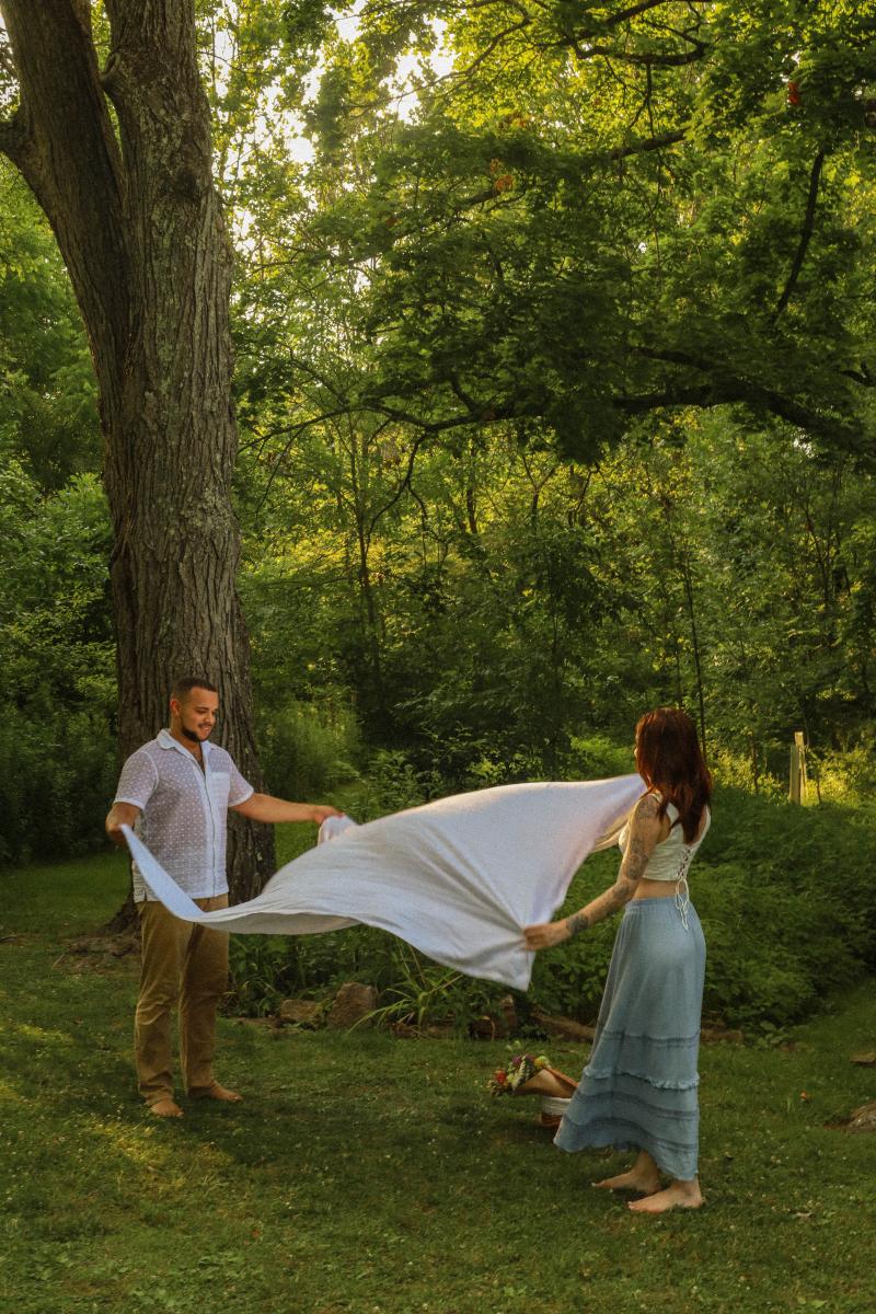 Couple spreading a picnic blanket under an old tree at the edge of the woods