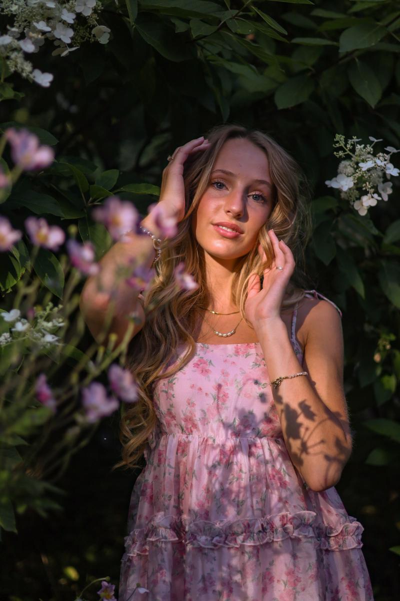 Waist-up outdoor portrait of a young woman in a floral dress, framed by pink and white blossoms in soft evening light