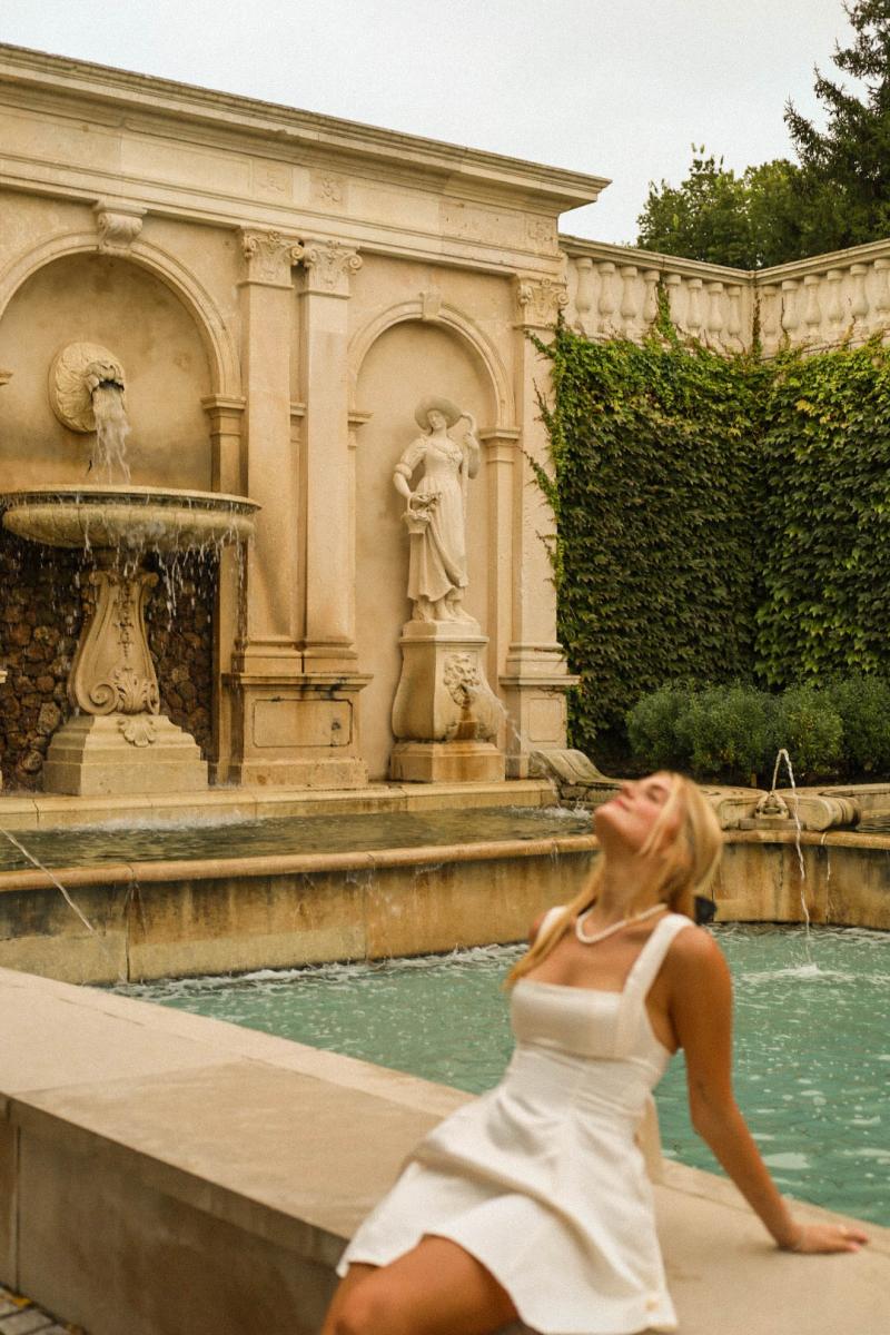 A young woman leaning back against the edge of a stone basin in front of a carved fountain wall