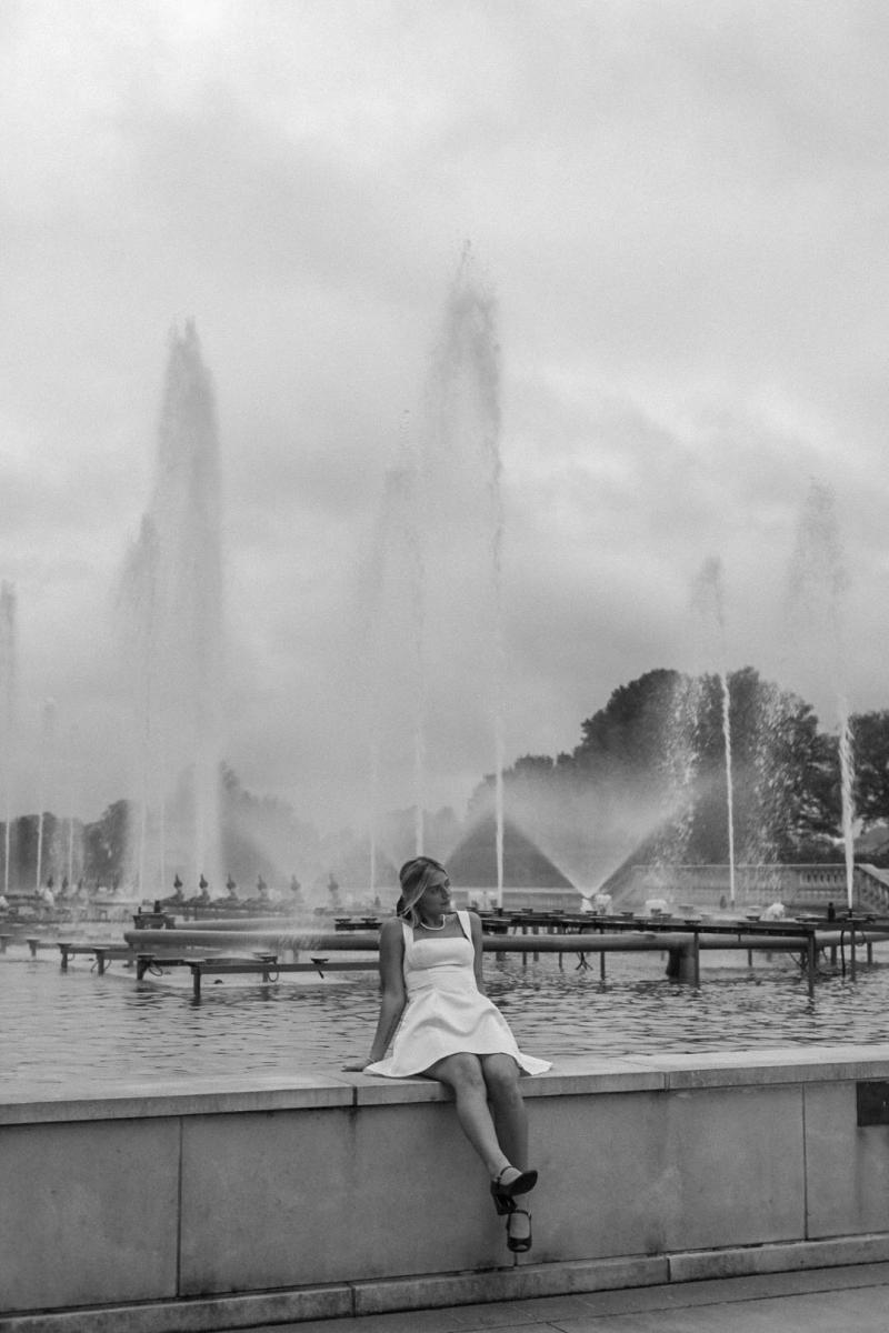 Black and white frame of a young woman sitting on a low stone wall in front of a long row of fountains