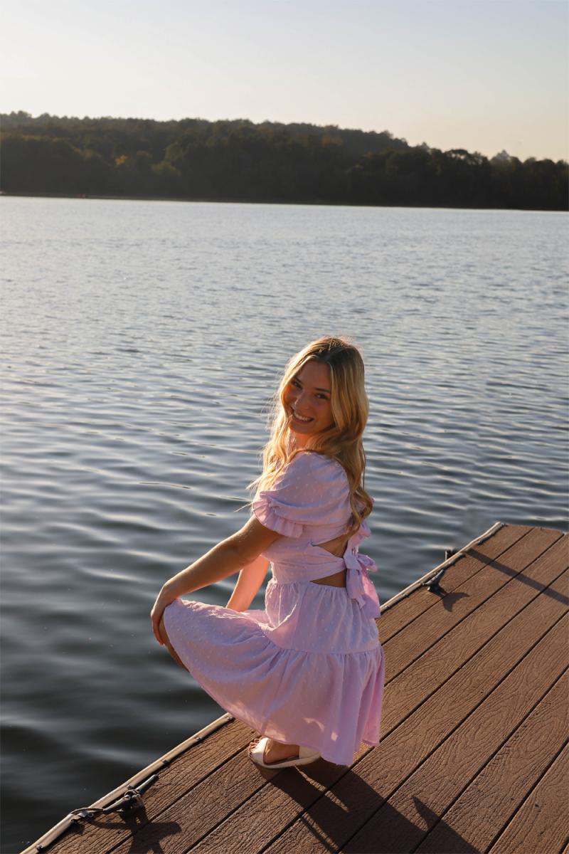 A young woman in a pink dress kneeling at the end of a wooden dock, open water and a wooded far bank behind her