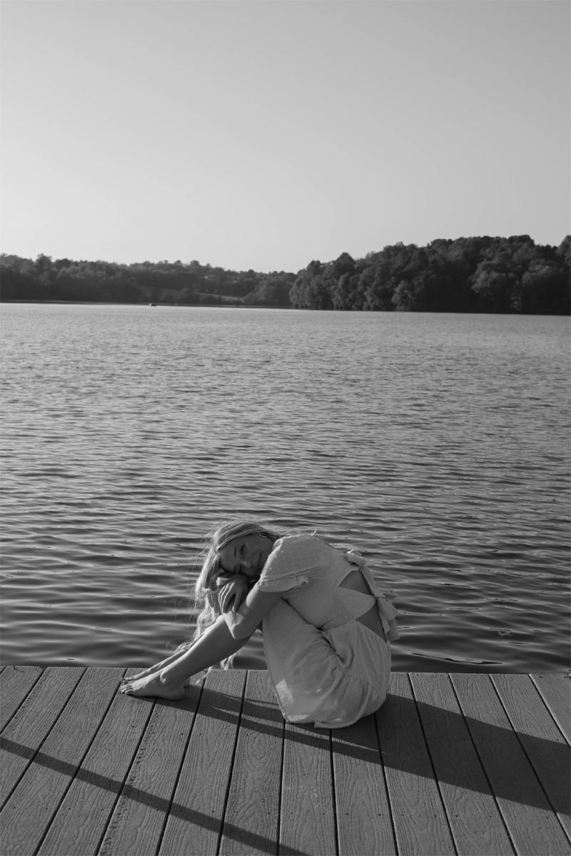 Black and white frame of a young woman sitting on a dock with her chin on her knees, water stretching to a far tree line
