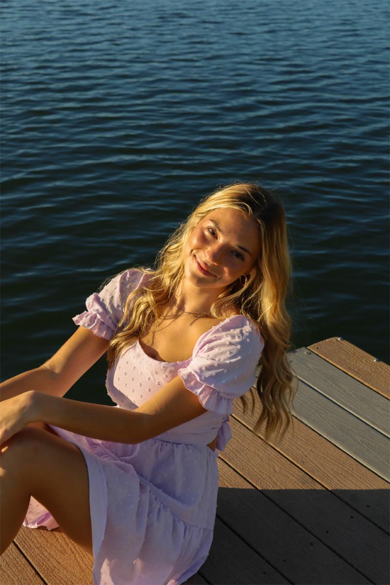 A young woman in a lilac dress sitting on a dock in late sun, deep blue water filling the frame behind her