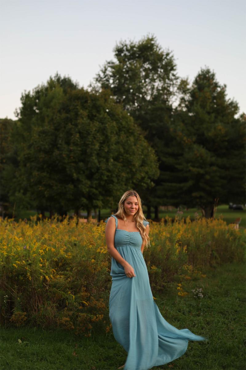 A senior in a long blue dress standing at the edge of a goldenrod field at dusk