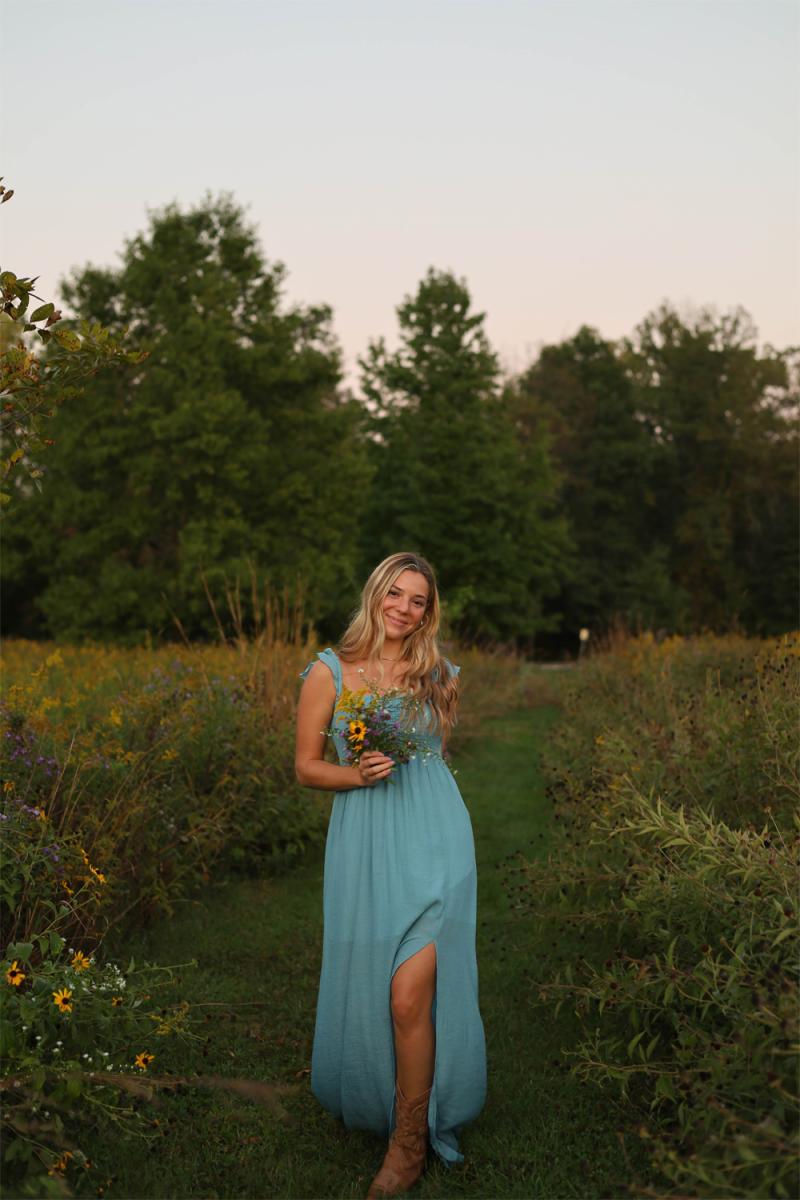A senior in a long blue dress walking a mown path through wildflowers, a small bunch of them in her hand