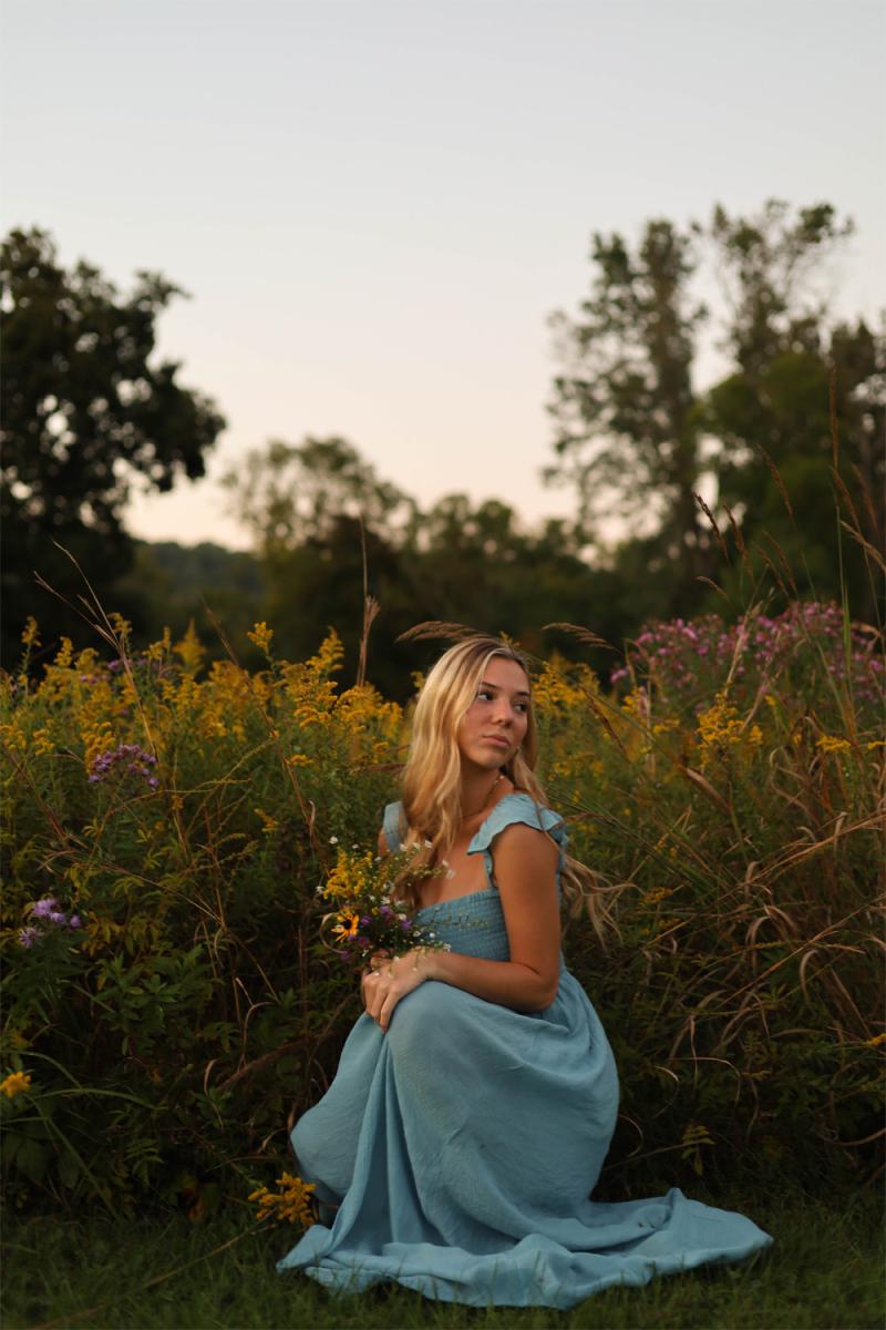 A senior kneeling in tall goldenrod, holding a small handful of wildflowers she has just picked