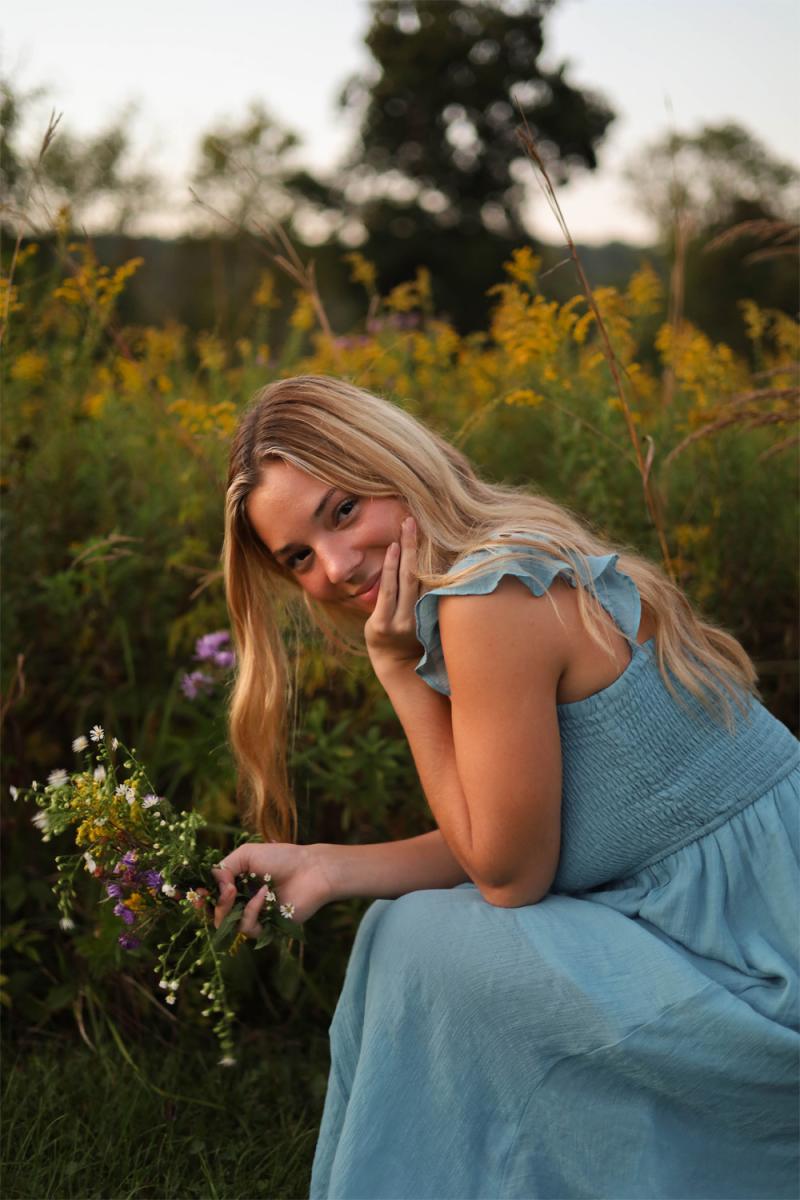 Outdoor portrait of a young woman in a blue dress resting her chin on her hand in a wildflower meadow at sunset