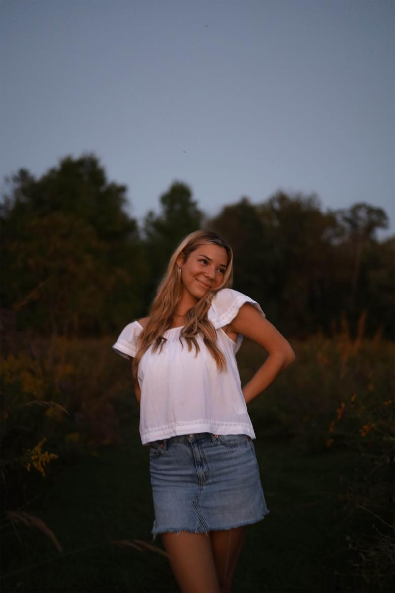 A senior in a white top and denim skirt at last light, the meadow behind her already going blue