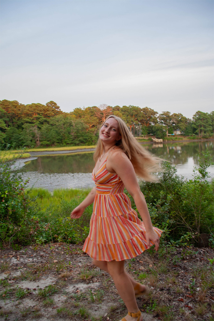 A young woman spinning in a striped dress on a bank above a quiet river bend, pines on the far side