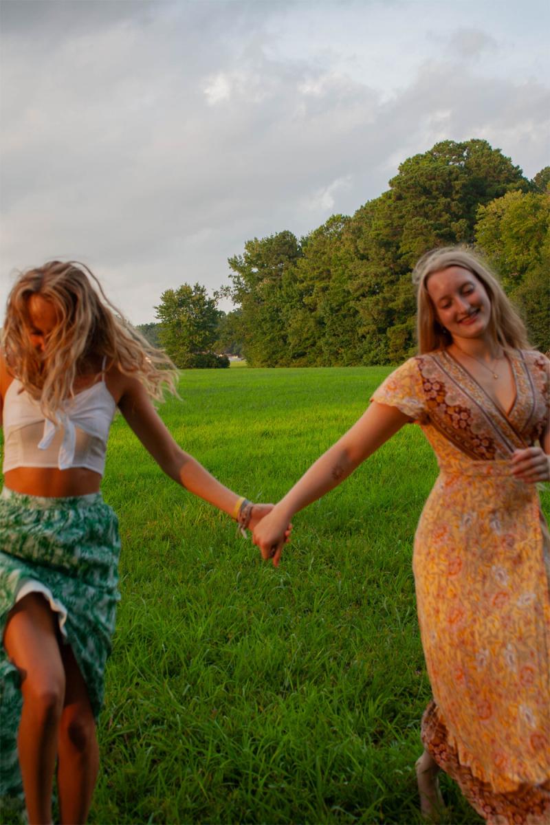 Two friends holding hands and walking away through a mown field, both of them laughing
