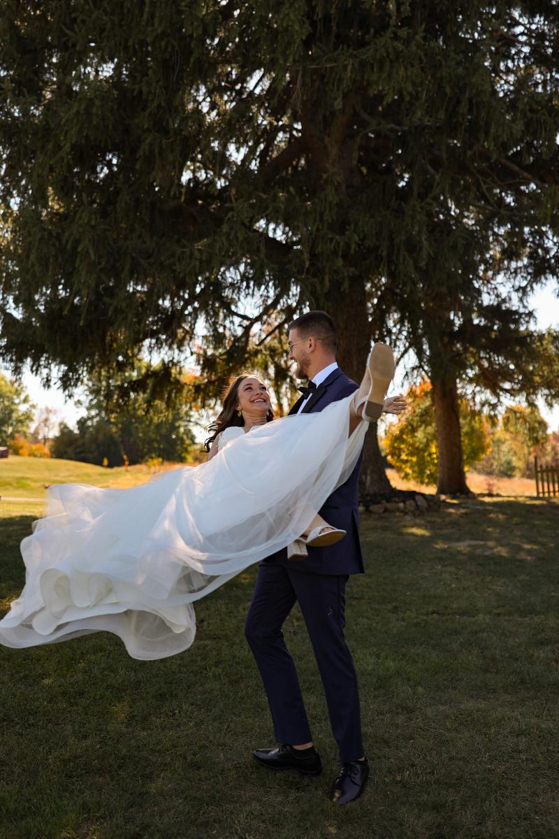 A groom in a navy suit swinging his bride off her feet under a big evergreen, her gown flying out behind her