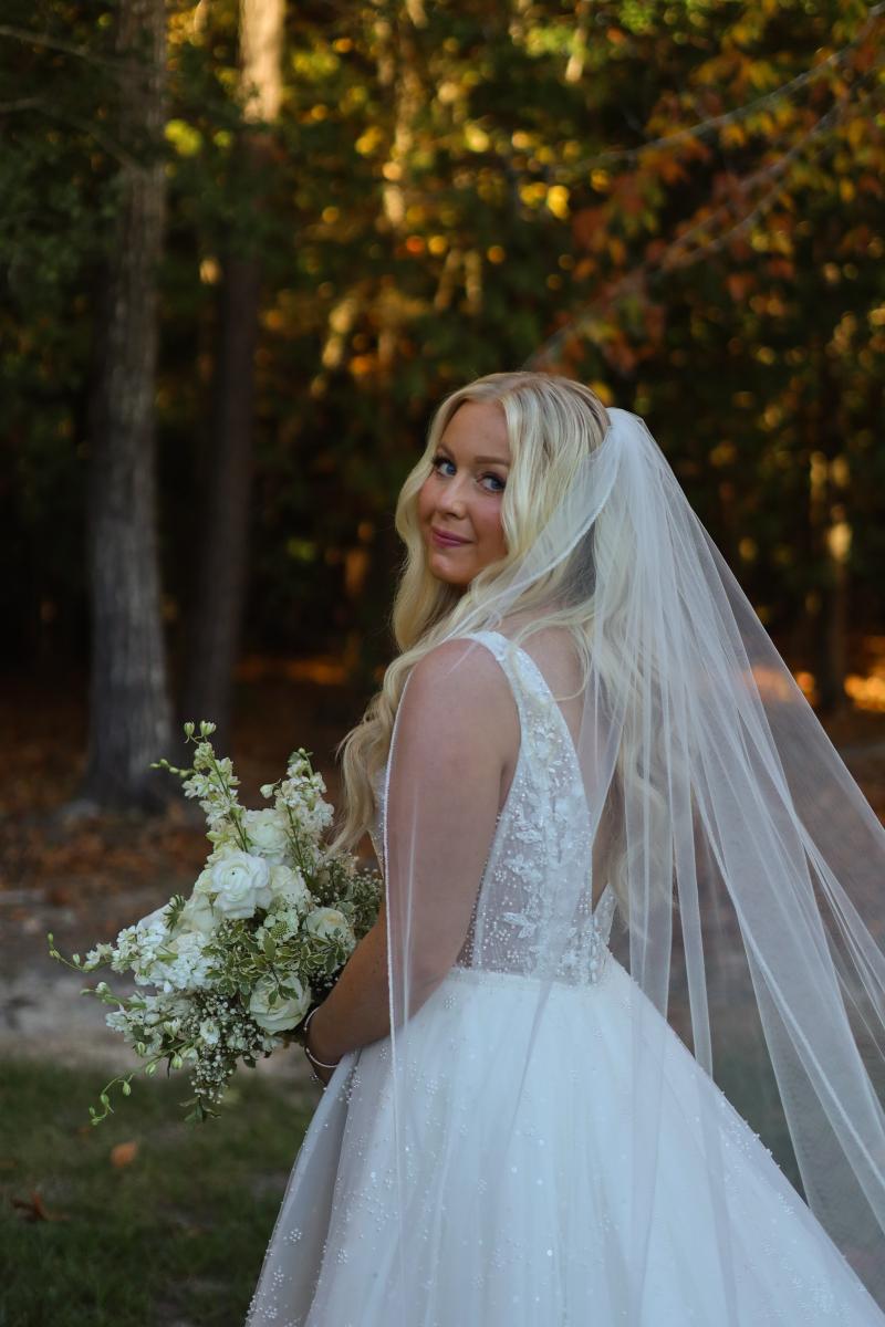 A bride looking back over her shoulder, a long veil trailing behind her and a white bouquet in her hand