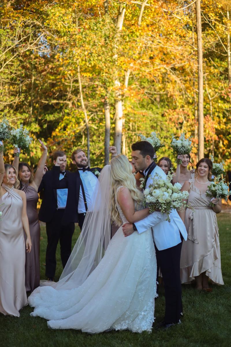 A bride and groom kissing while their wedding party cheers and lifts their bouquets around them