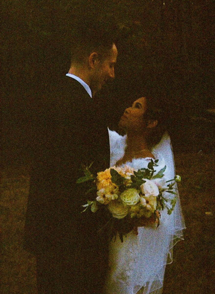 A bride and groom standing close together after dark, her bouquet held between them, lit by one warm light